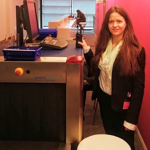 Sicherheitsmitarbeiterin steht neben einem Gepäckscanner an einem Kontrollpunkt und hält ein Funkgerät in der Hand